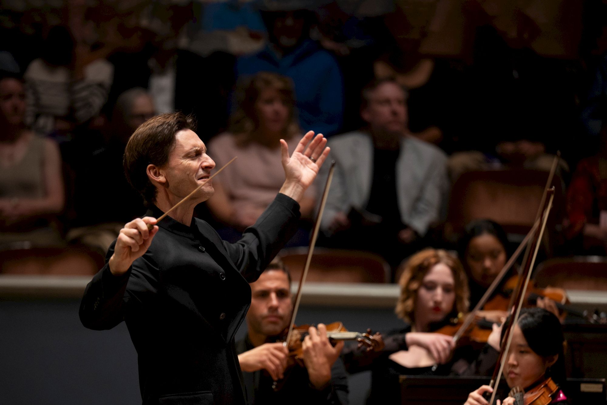 A female conductor leads an orchestra, baton raised, with musicians and an audience behind her, focused on the performance.