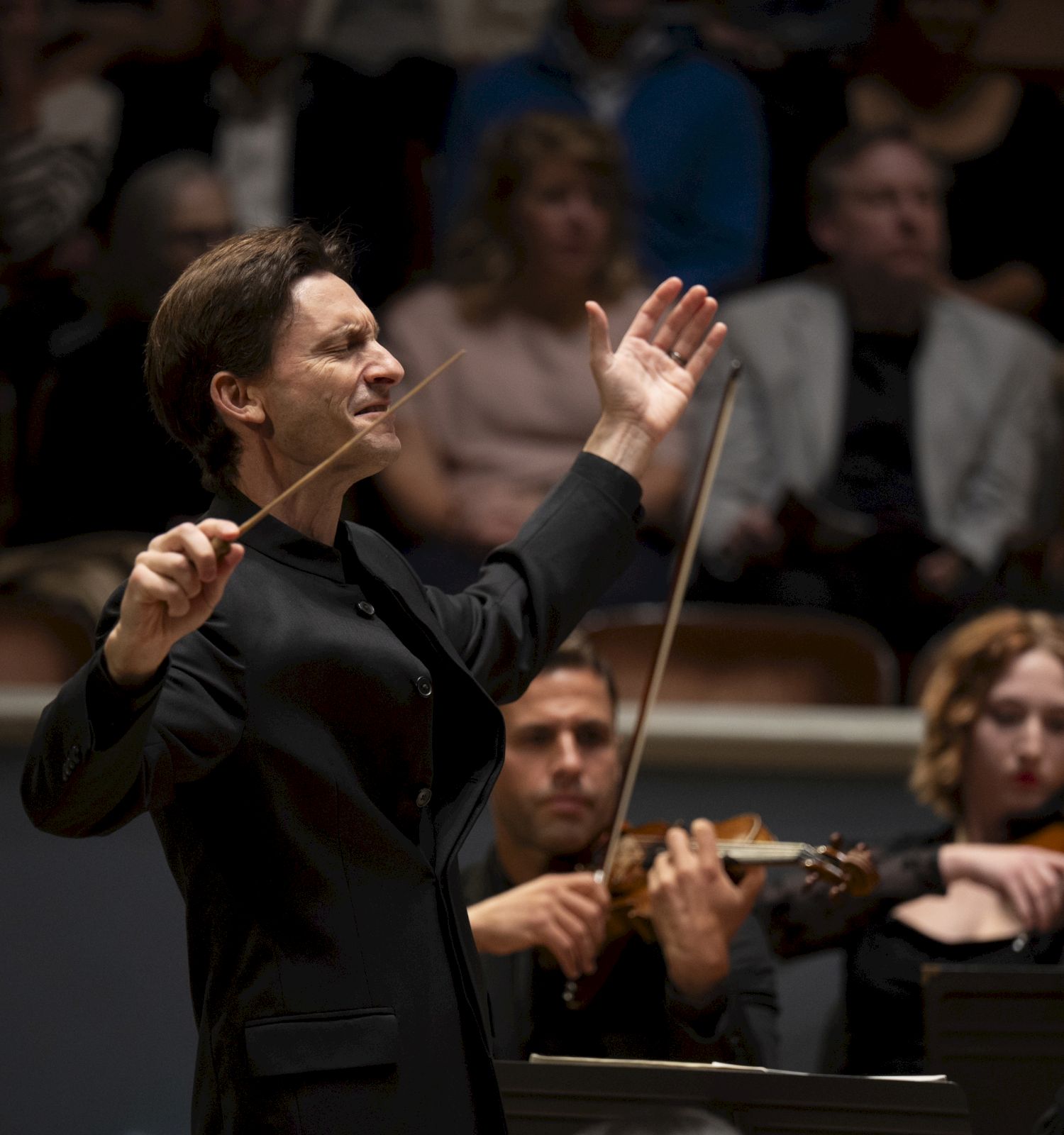 A conductor leads an orchestra with a baton, surrounded by musicians playing strings, in a concert hall with an attentive audience.