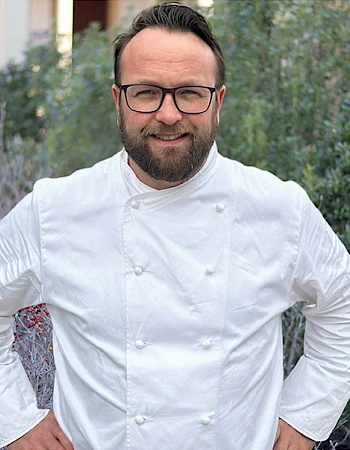 A man in a white chef&rsquo;s jacket stands outdoors with hands on hips, glasses, trimmed beard, greenery and a building in the background.