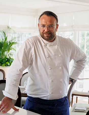 A chef in a white jacket stands by a counter in a bright, modern kitchen, looking at the camera with a slight smile.