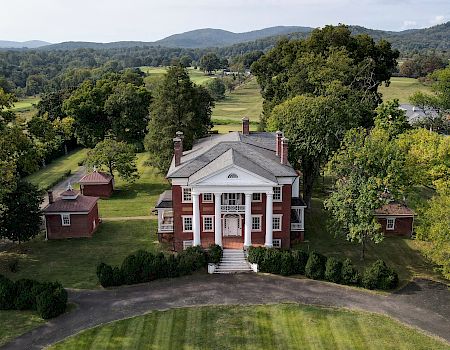 A large, historic white-columned mansion with a red brick base, surrounded by green lawns, trees, and small outbuildings in a rural setting.
