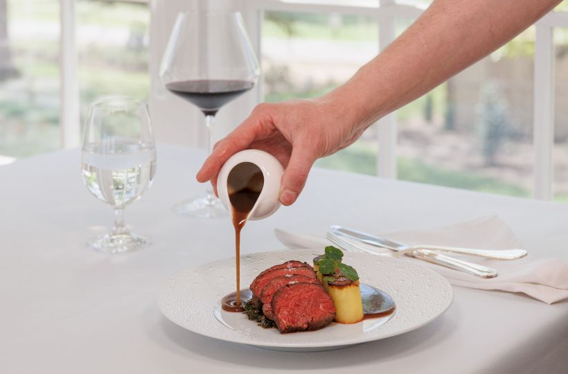 A chef pours a rich sauce over a plated beef dish with garnish and wine in the background, on a fine dining table.