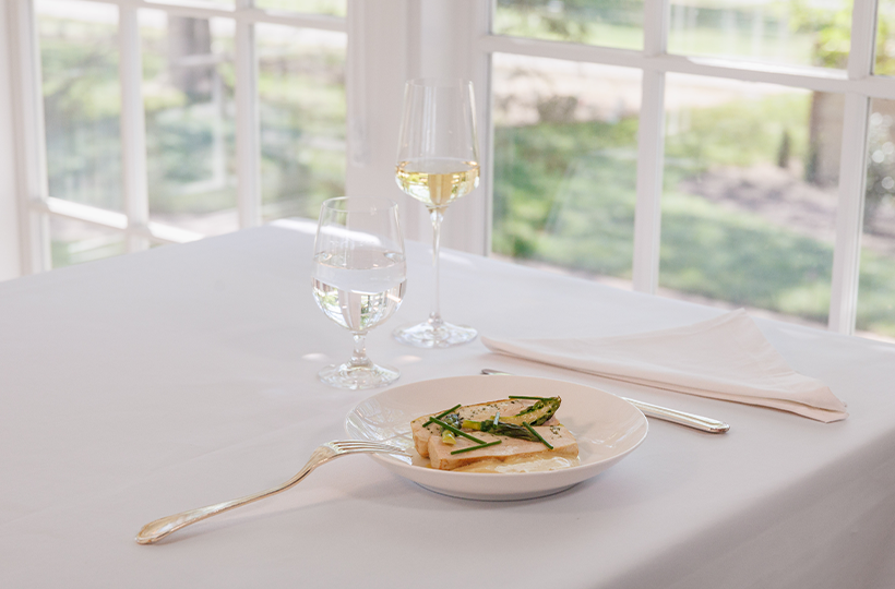 A single plate with toasted bread topped by herbs sits on a white table, flanked by a wine glass and a water glass, near a napkin.