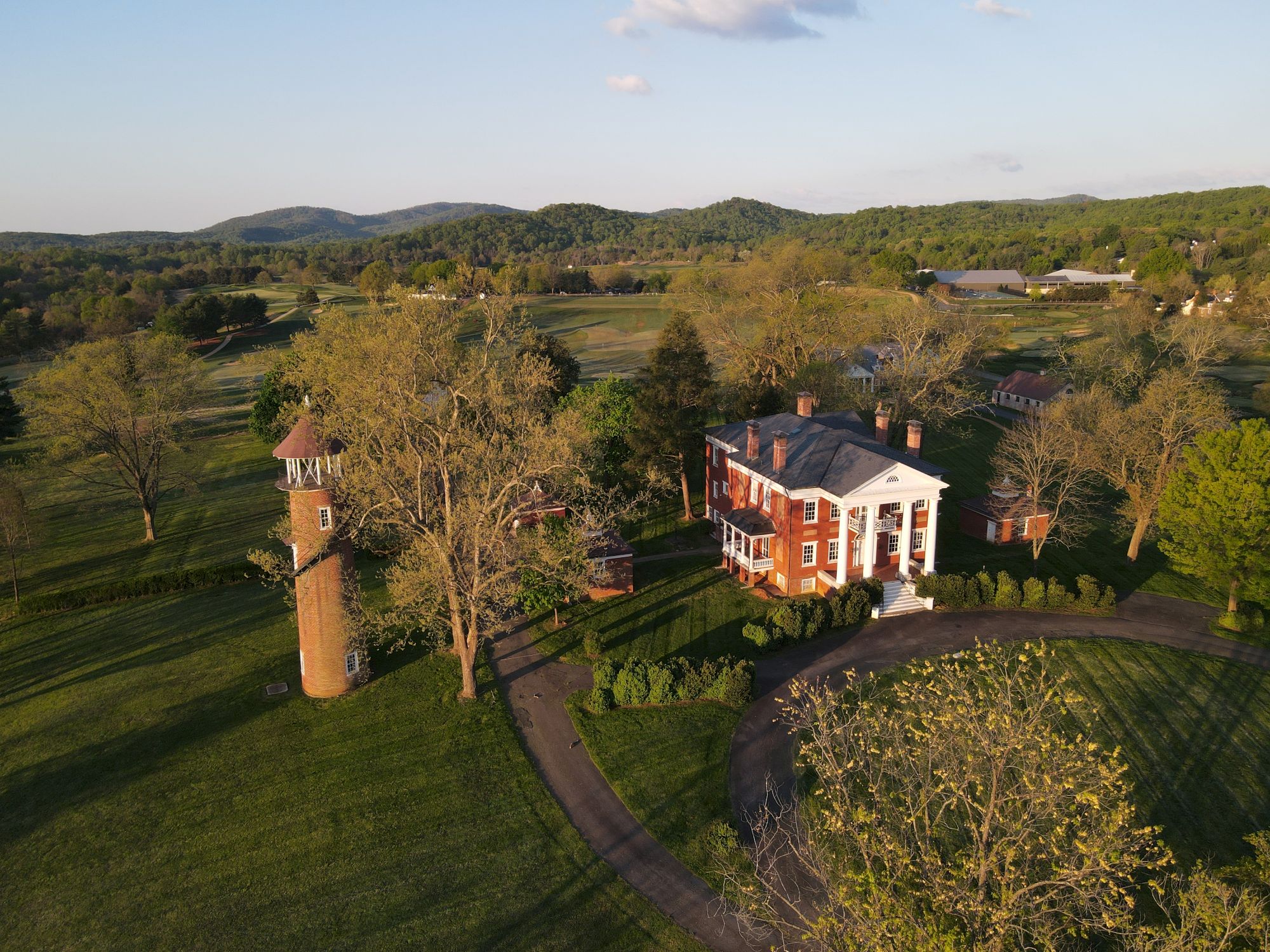 Aerial view of a brick building with a white house, surrounded by trees, a curved driveway, and a tall brick water tower on a green campus-like landscape.