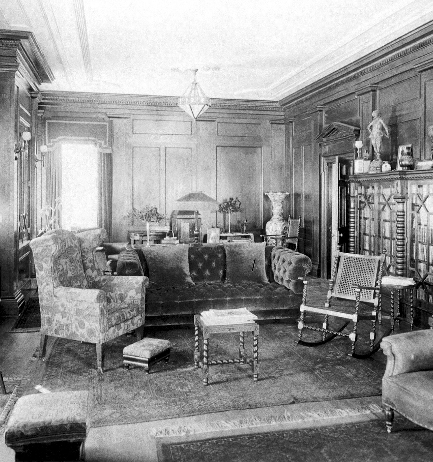 A vintage living room with wood paneling, cushioned chairs, a fireplace, and a mix of seating around a central rug, giving an old-world parlor vibe.