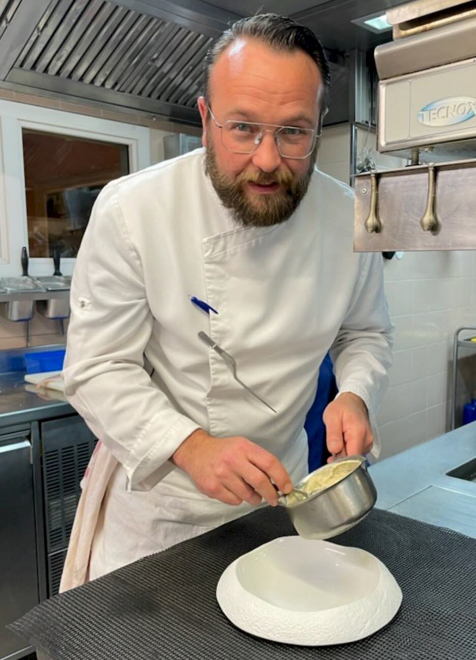 A chef in a white coat pours or sauces into a small bowl over a white plate, focused in a bustling kitchen.
