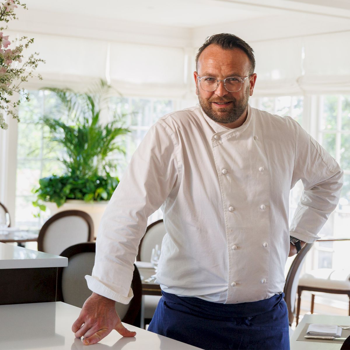 A chef in a white coat stands by a kitchen island, looking toward the camera with one hand resting on the counter.