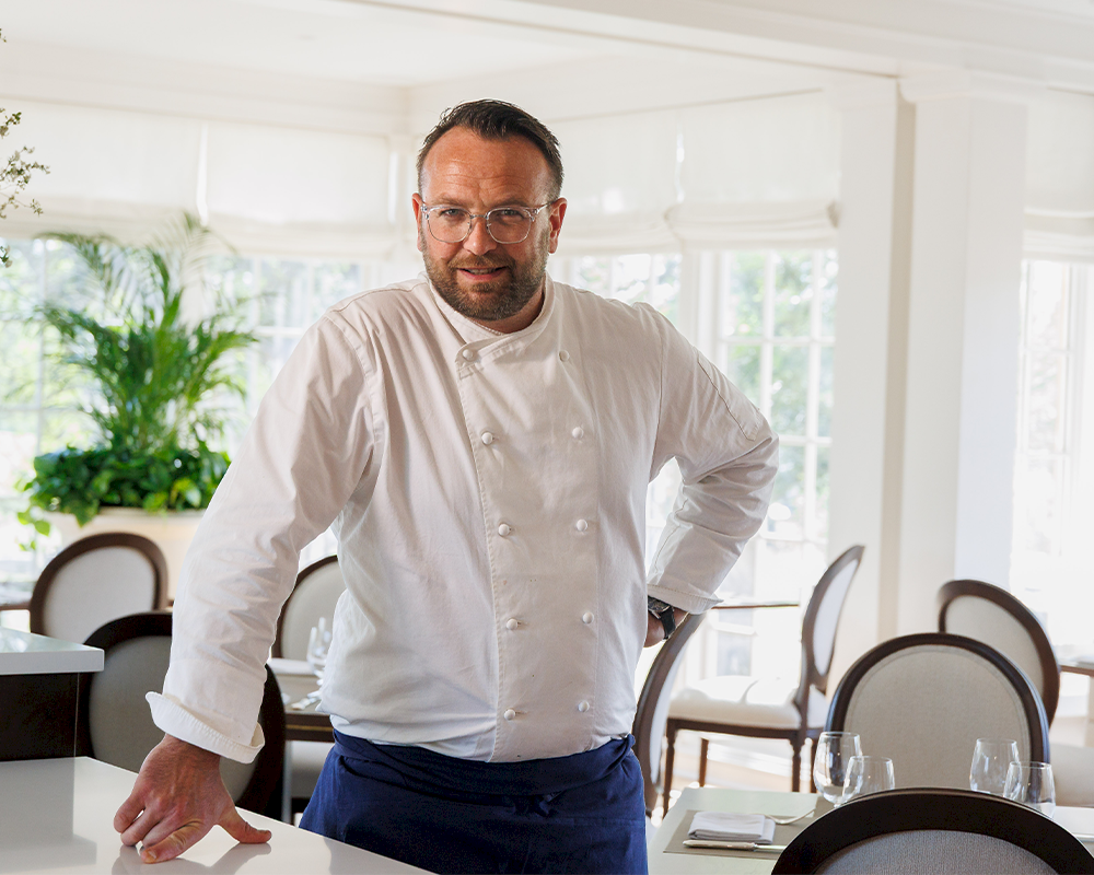 A chef in a white jacket stands by a table in a bright, airy dining area with plants and empty chairs, ready to serve.