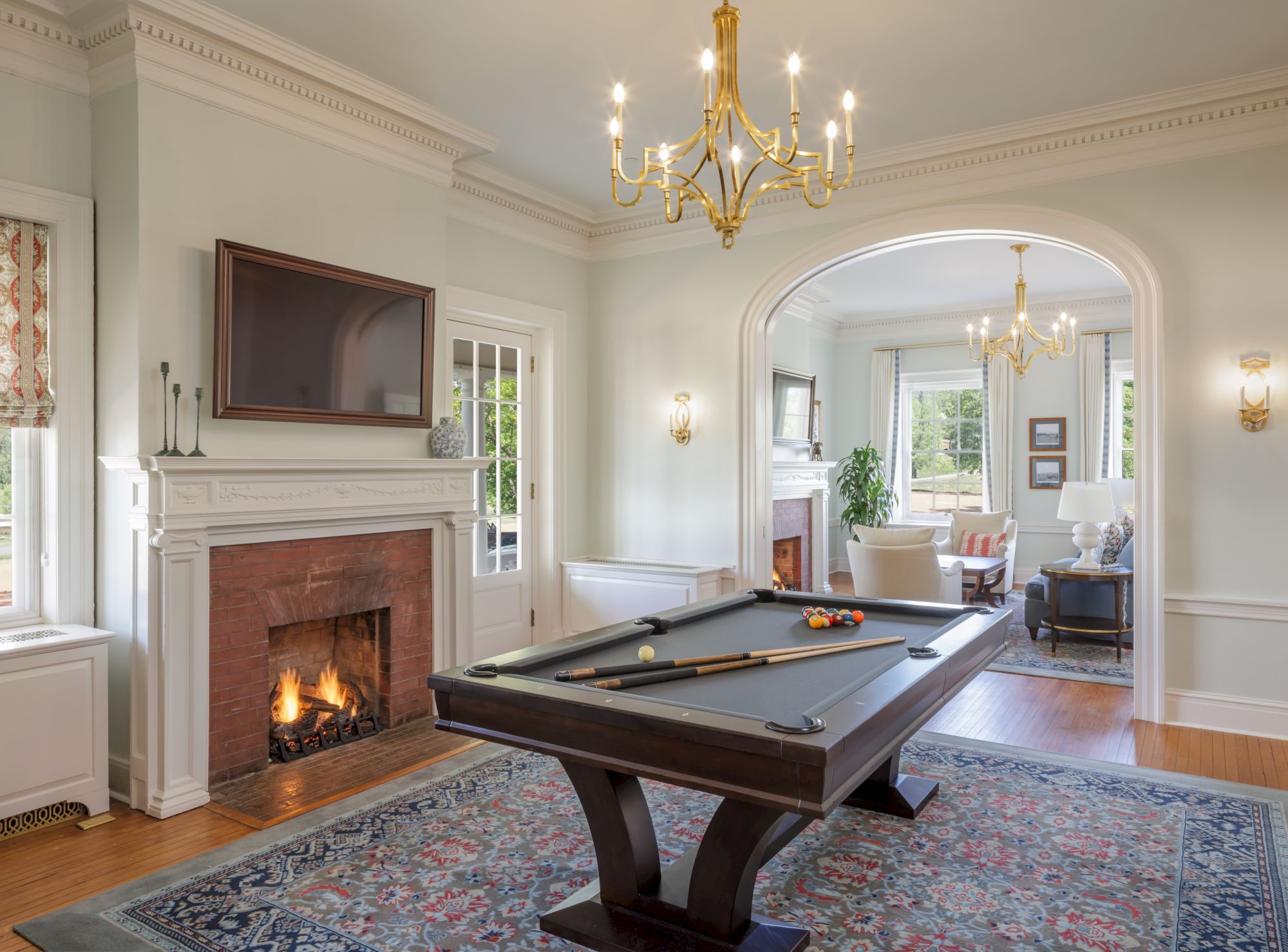 A luxurious game room with a dark pool table, fireplace, chandeliers, and a connected living area visible through an arched doorway.