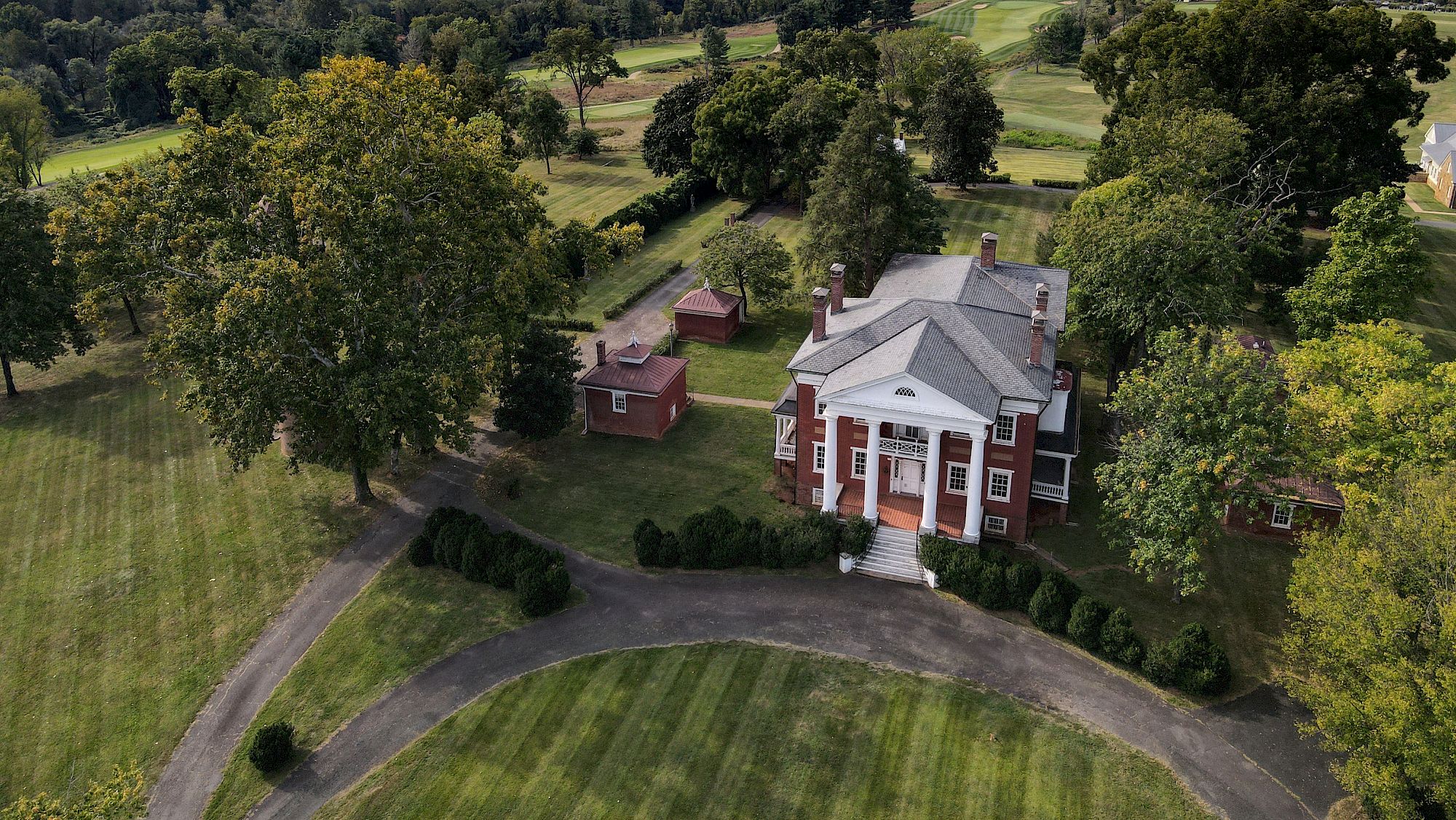 An aerial view of a stately brick mansion with a white portico, curved driveway, surrounding green lawns, and mature trees.