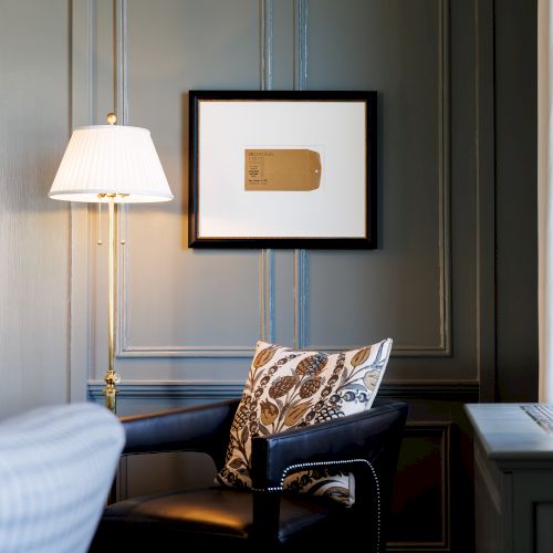 A cozy, stylish hotel reading nook with a dark paneled wall, framed art, a lamp, a patterned cushion, and a small table by sheer curtains.