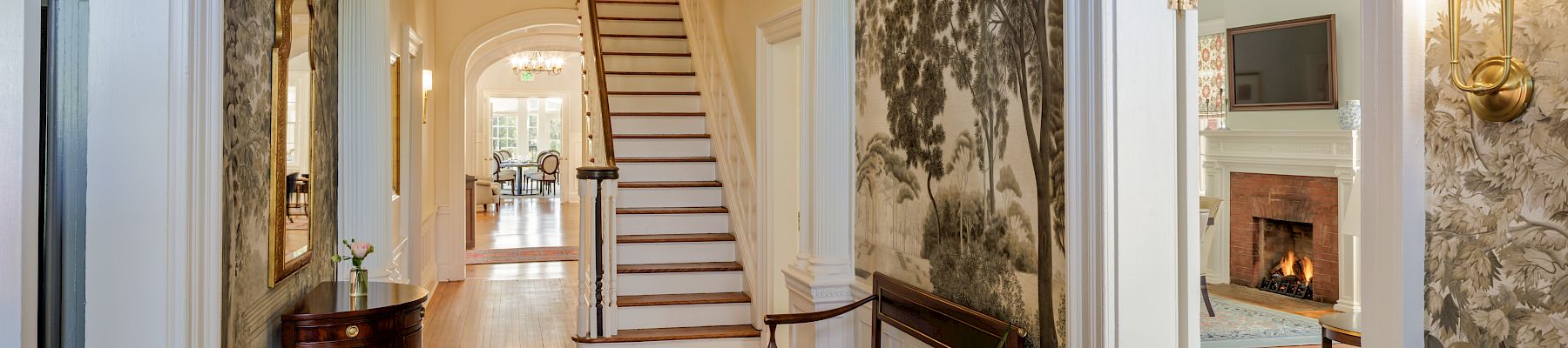 A grand hallway with an arched doorway, staircase at the end, vintage wallpaper, hardwood floors, a rug, potted plant, and a mirror reflecting a room with a fireplace.