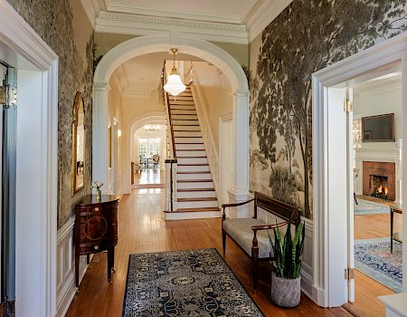 A grand hallway with an arched doorway, staircase at the end, vintage wallpaper, hardwood floors, a rug, potted plant, and a mirror reflecting a room with a fireplace.