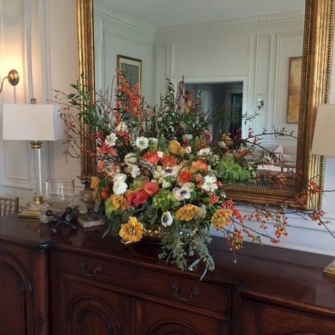 A large ornate floral arrangement sits atop a dark wooden sideboard beneath a gilded mirror, flanked by two gold lamps in a classic, elegant room.