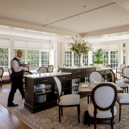 A bright, elegant dining area with round-backed chairs, a long central counter, and large windows; a staff member tends the space, inviting a refined ambiance.