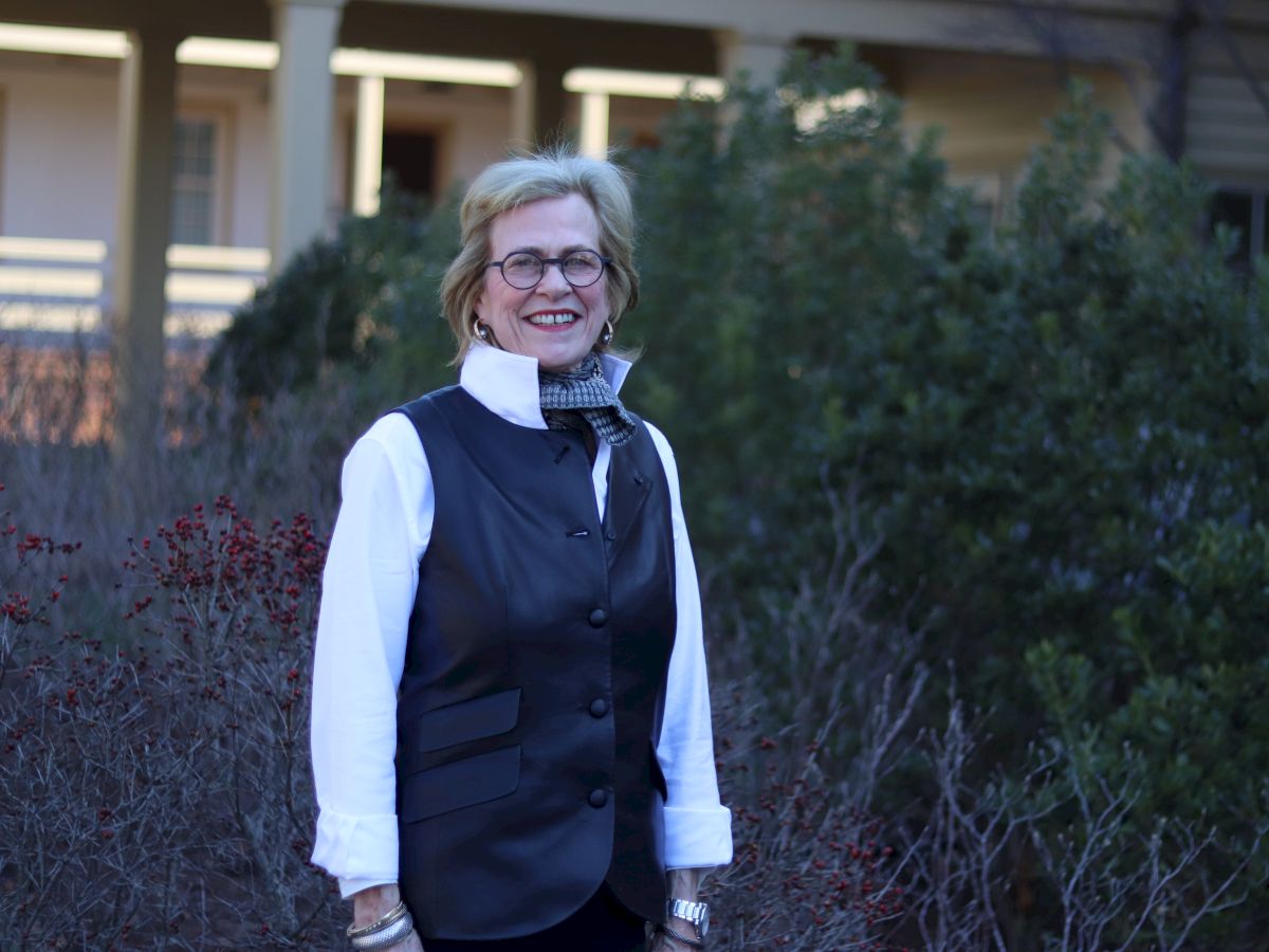 A smiling woman stands outdoors in a white blouse and black vest, with glasses, in front of shrubs and a building.