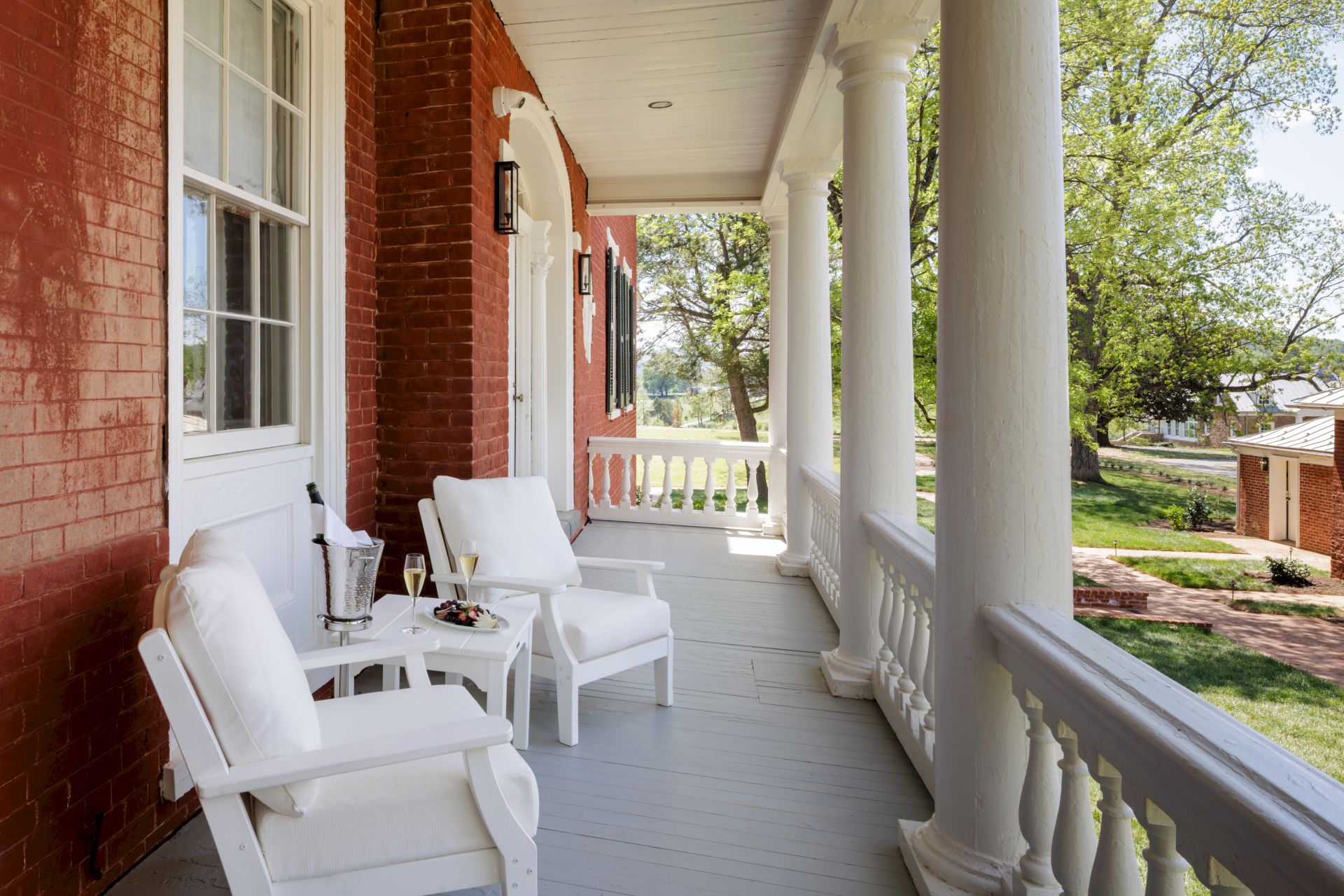 A sunny porch with white wooden chairs and a small table along a brick house, tall columns, and a green yard in the background.