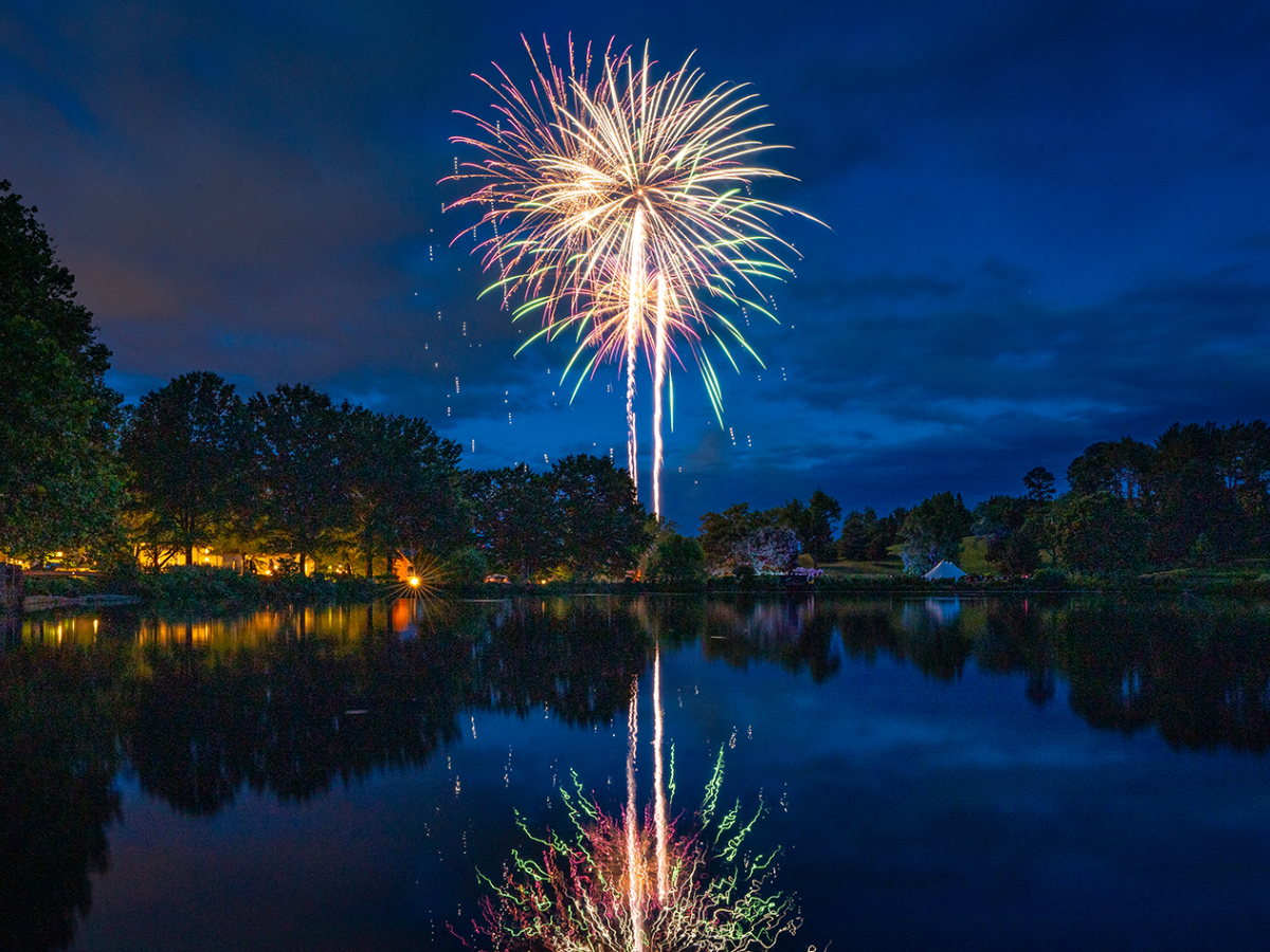 Fireworks explode above a calm lake at night, casting colorful reflections on the water, with trees and a distant shoreline lit by lights.