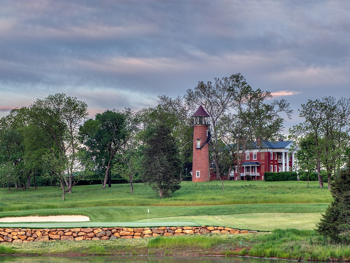 A green golf course with sand bunker, a red brick tower/lighthouse and mansion, trees, and a bright moon at dusk.