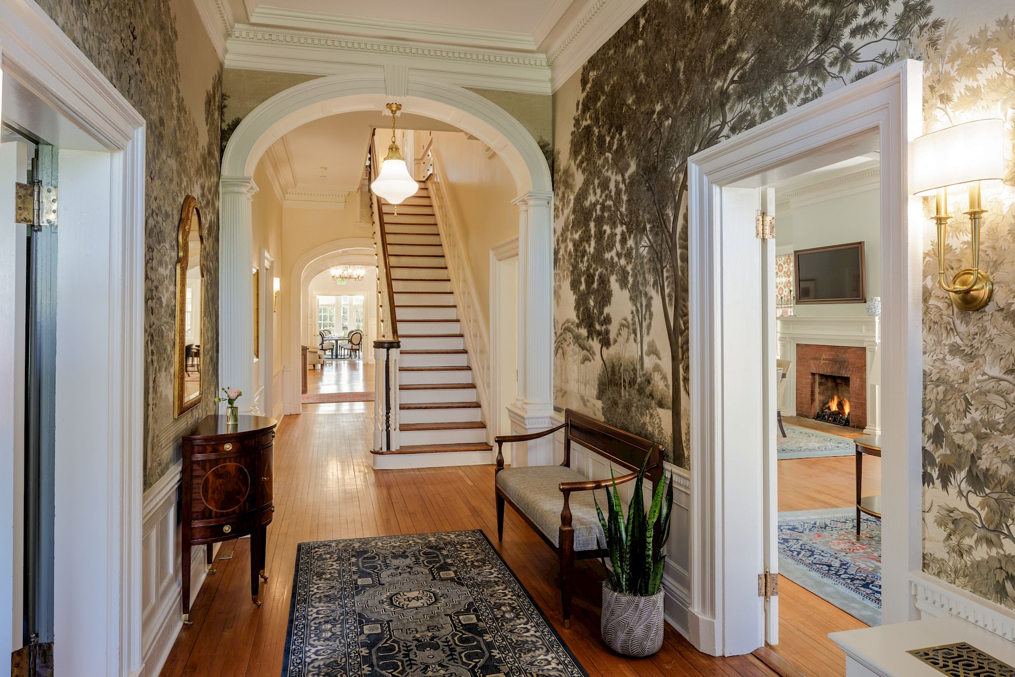 A grand foyer with a staircase, arched entry, chandelier, patterned wallpaper, wooden floors, a rug, a plant pot, and a doorway to a living room with a fireplace.