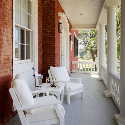 A porch with white chairs, a small table, and a brick house, featuring columns and a green yard beyond, sunny and inviting.