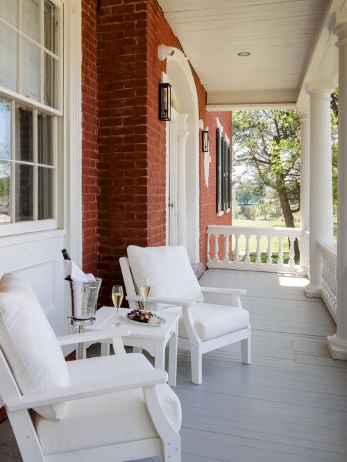 A porch with white chairs, a small table, and a brick house, featuring columns and a green yard beyond, sunny and inviting.