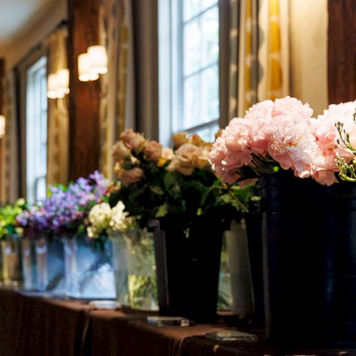 A row of colorful bouquets in pots lined along a table against a wall with windows, creating a bright, floral display.