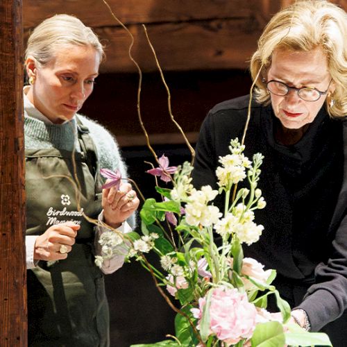 Two women arrange a floral bouquet indoors, with one wearing an apron and the other in glasses, focusing on the flowers.
