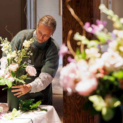 A florist arranges a lush flower bouquet on a table, leaning over with care, blurred colorful blooms nearby.