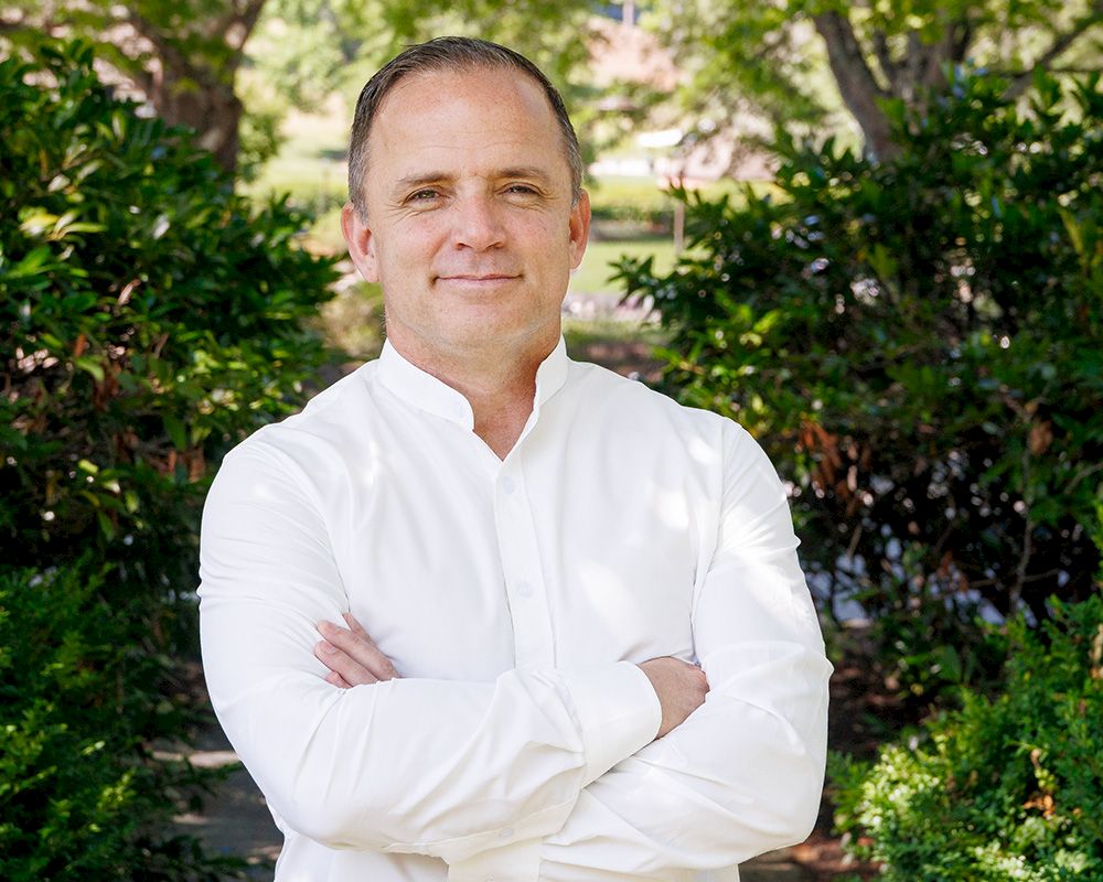 A man in a white shirt stands outdoors with arms crossed, in front of green shrubs and trees, a calm, confident pose.