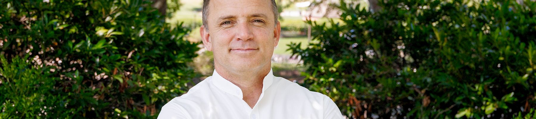 A man in a white button-down shirt stands with arms crossed, outdoors, in front of green bushes and trees on a sunny day.