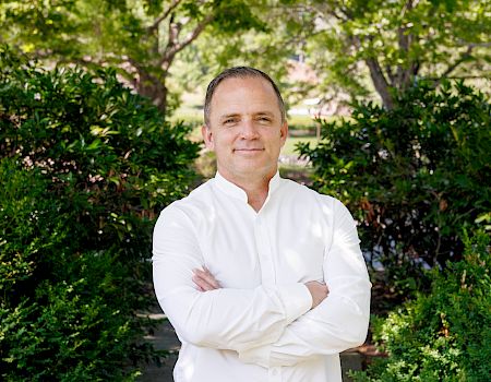 A man in a white button-down shirt stands with arms crossed, outdoors, in front of green bushes and trees on a sunny day.