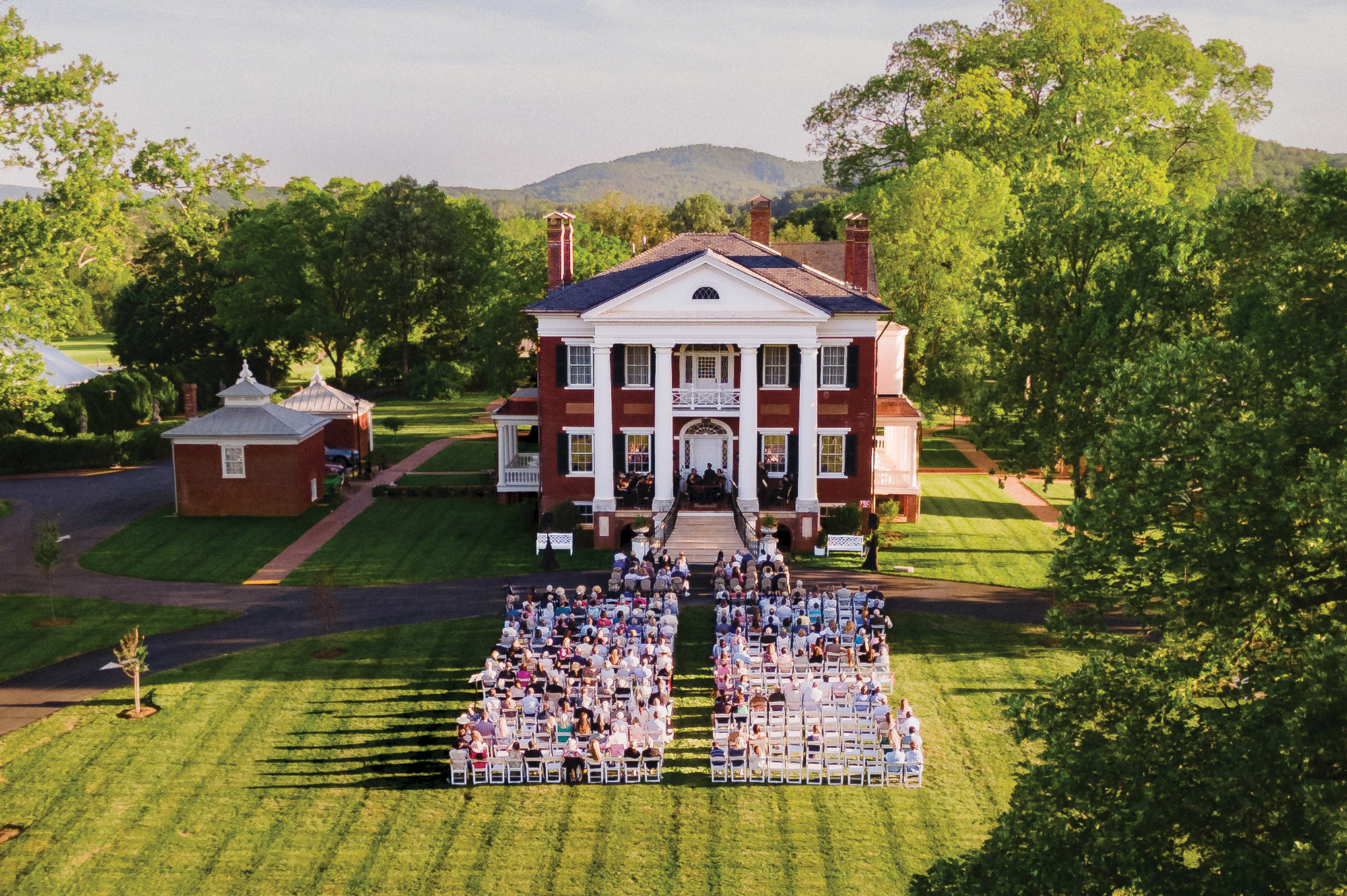 An aerial view of a white-columned campus building with a large outdoor wedding ceremony on the lawn, guests seated in rows, sunny day.