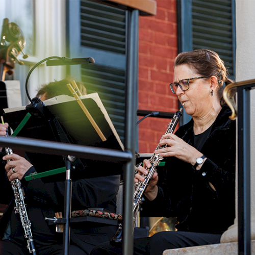 A small ensemble outdoors plays woodwind instruments, including clarinets, with music stands and a guitarist visible in the background, as patrons listen.