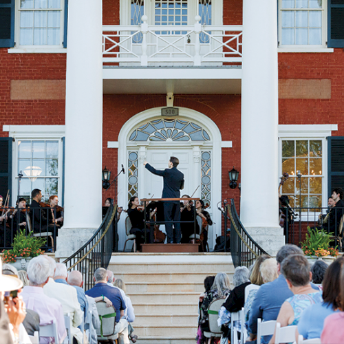 A brass band performs on steps of a red brick building with white columns, while seated guests watch a ceremony on a sunny day.
