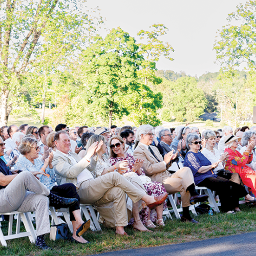 A crowd of people seated outdoors on white chairs, watching an event in a sunny park with green trees and grass.