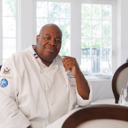A bald chef in a white jacket sits at a restaurant table, smiling thoughtfully in front of large windows.