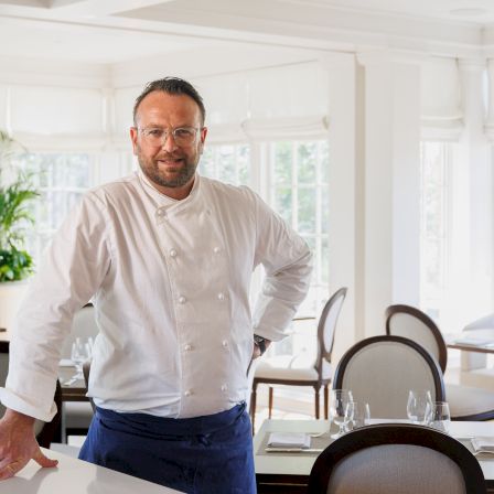 A chef in a white jacket smiles at a bright restaurant table with chairs, glasses, and large windows behind him.