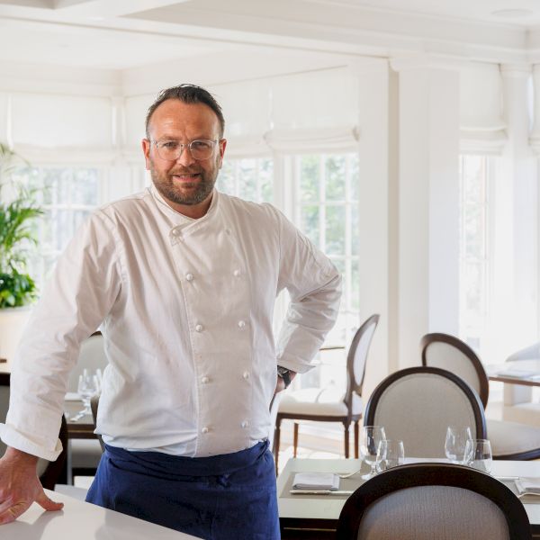 A chef in a white jacket smiles at a bright restaurant table with chairs, glasses, and large windows behind him.