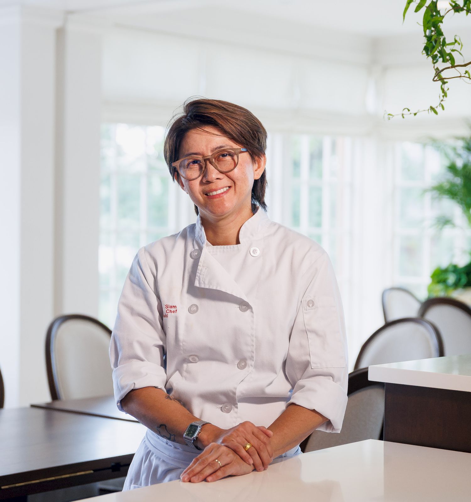 A smiling female chef in a white jacket and glasses sits at a table in a bright restaurant with plants.