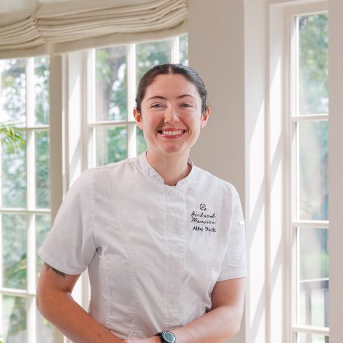 Smiling chef Abby stands indoor The Dining Room, with plants around, radiating a welcoming, professional vibe.