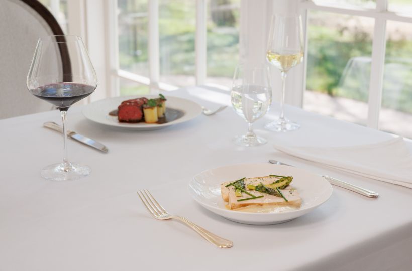 A fine-dining table with white plates, plated dishes, and two wine glasses in front of bright windows.