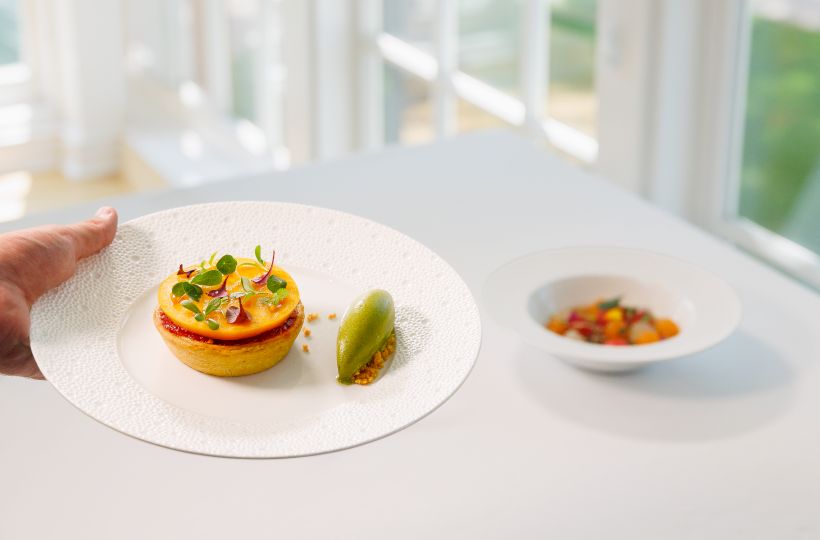 A server holds a white plate with a garnished mini tart and a green stuffed bite, with another dish blurred behind.