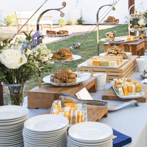 Outdoor buffet table at a garden party with pastries, sandwiches, and cake slices served on tiered stands.