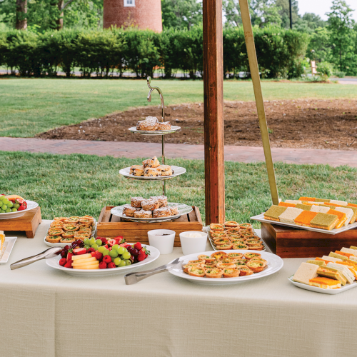 An outdoor buffet with cakes, pastries, fruit, and sandwiches on stands in a green park yard under a pavilion.