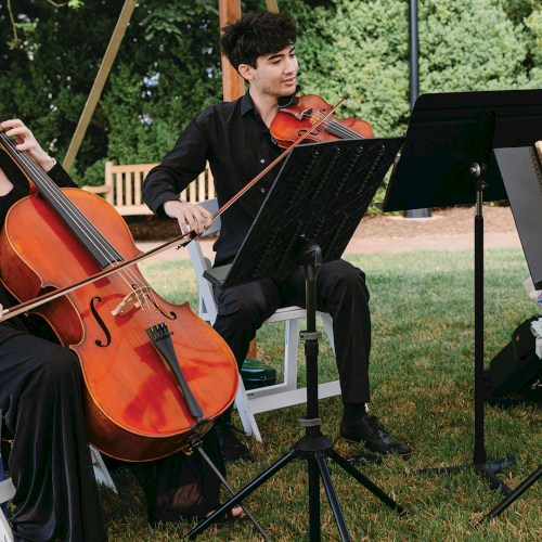 An outdoor string ensemble plays cello and violin on folding chairs with a music stand and microphone, surrounded by trees.