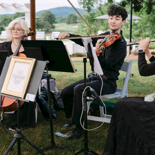 An outdoor trio performs violin, viola, and cello on folding chairs in a garden-like venue.