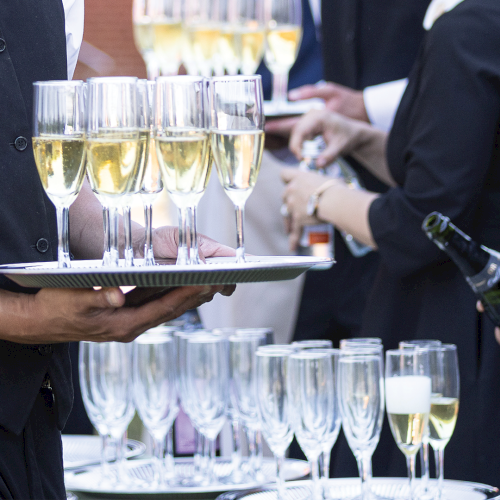 A server holds a tray of champagne flutes at a formal event as guests prepare to toast, clinking their sparkling drinks.