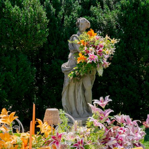 A garden statue holding a colorful bouquet stands behind a table set with bright lilies, other flowers, candles, and utensils.