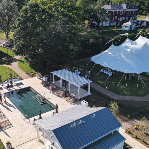 An aerial view of a backyard event with a large white tent, guests, and a blue swimming pool in the courtyard.